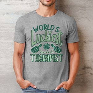 May include: A gray t-shirt with the text "WORLD'S Luckiest Speech Therapist" in green. The design includes a shamrock and two thumbs-up symbols. The shirt is made of a soft material.