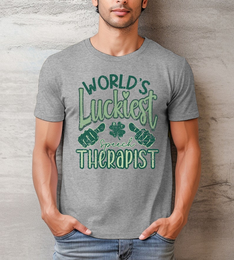 May include: A gray t-shirt with the text "WORLD'S Luckiest Speech Therapist" in green. The design includes a shamrock and two thumbs-up symbols. The shirt is made of a soft material.