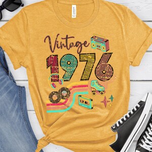 May include: Mustard yellow t-shirt with the text "Vintage 1976" and retro graphics including a boombox, roller skate, cassette tape, and records. The shirt has a crew neck and short sleeves.