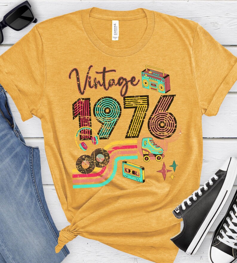May include: Mustard yellow t-shirt with the text "Vintage 1976" and retro graphics including a boombox, roller skate, cassette tape, and records. The shirt has a crew neck and short sleeves.