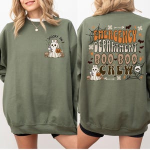 May include: Olive green sweatshirt with a Halloween-themed design. The front features a ghost wearing a nurse's cap, holding a syringe and a pumpkin. The back displays the text "Emergency Department Boo-Boo Crew" with Halloween graphics.