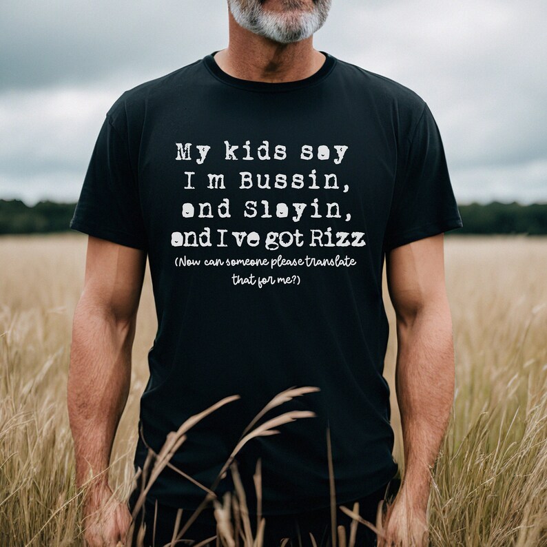 May include: Black t-shirt with white text that reads "My kids say I'm Bussin, and Slayin, and I've got Rizz (Now can someone please translate that for me?)". The shirt is worn by a person standing in a field.