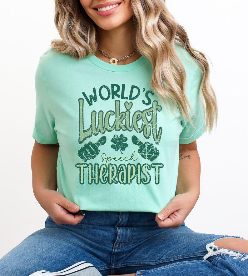 May include: A light green t-shirt with the text "WORLD'S Luckiest Speech Therapist" in distressed green lettering. The design includes a shamrock and hand gestures. The shirt is made of a soft material.