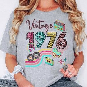 May include: A gray t-shirt with the text "Vintage 1976" in a retro design. The shirt features illustrations of a boombox, roller skate, cassette tape, and records, with colorful stripes and star accents.