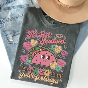 May include: A gray t-shirt with a pink and green taco graphic and the text "Tis the Season to Taco 'bout your feelings". The taco has a smiling face and is surrounded by pink hearts with text like "Feel the Feels", "You Matter", and "Let's Talk".