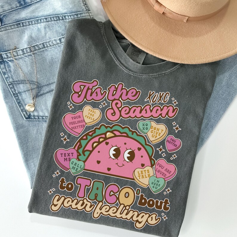 May include: A gray t-shirt with a pink and green taco graphic and the text "Tis the Season to Taco 'bout your feelings". The taco has a smiling face and is surrounded by pink hearts with text like "Feel the Feels", "You Matter", and "Let's Talk".