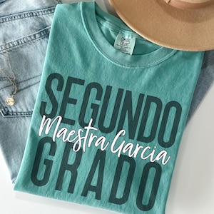 May include: A teal Comfort Colors t-shirt with the words "SEGUNDO Maestra Garcia GRADO" printed in dark teal and white. The shirt is folded and placed on top of a pair of light blue jeans. A gold necklace and a tan hat are also visible.