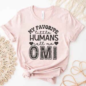 May include: A pink t-shirt with the text "My favorite little humans call me OMI" in black with a distressed font.