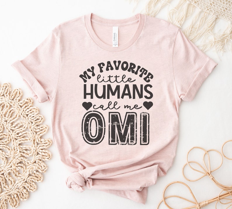 May include: A pink t-shirt with the text "My favorite little humans call me OMI" in black with a distressed font.