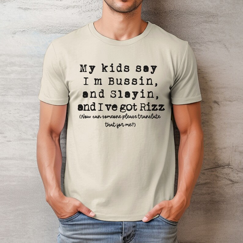 May include: A light beige t-shirt with black text that reads "My kids say I'm Bussin, and Slayin, and I've got Rizz (Now can someone please translate that for me?)". The shirt is short-sleeved.