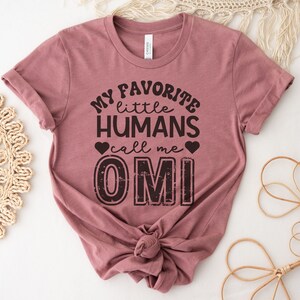 May include: A mauve t-shirt with the text "My favorite little humans call me OMI" in black with a distressed font. The text is centered on the shirt.
