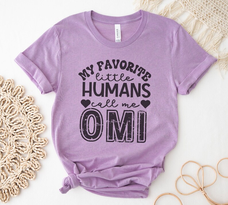 May include: A lavender t-shirt with the text "My favorite little humans call me Omi" in black. The text is in a distressed font and has a heart on either side of the word "call".