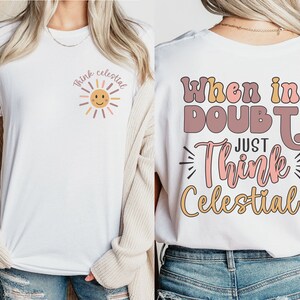 Puede incluir: Camiseta blanca con un gr&aacute;fico de sol rosa y amarillo y el texto "Think Celestial" en la parte delantera. La parte posterior de la camiseta tiene el texto "When in Doubt Just Think Celestial!" en rosa, amarillo y marr&oacute;n.