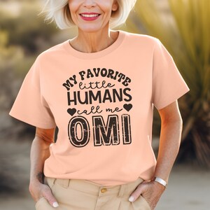 May include: A peach-colored t-shirt with black text that reads "My favorite little humans call me Omi".