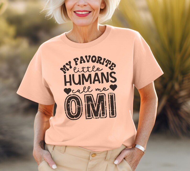 May include: A peach-colored t-shirt with black text that reads "My favorite little humans call me Omi".