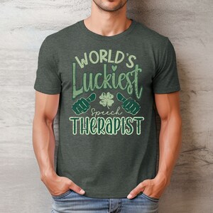 May include: Dark gray t-shirt with the text "WORLD'S Luckiest Speech Therapist" in green. The design includes a four-leaf clover and thumbs-up symbols. The shirt is made of a soft material and has a crew neck.