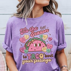 May include: Purple t-shirt with a graphic of a taco with a smiling face. The taco is surrounded by candy hearts with messages like "Feel the Feels", "You Matter", "Don't Give Up", "Let's Talk", "You Are Loved", and "I'm Here 4 U". The text on the shirt reads "Tis the Season to Taco'bout your feelings".