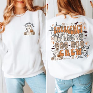 May include: White sweatshirt with a Halloween-themed design. The front features a ghost nurse with the text "Spooky RN." The back displays "Emergency Department Boo-Boo Crew" with bats, cobwebs, and a skull.