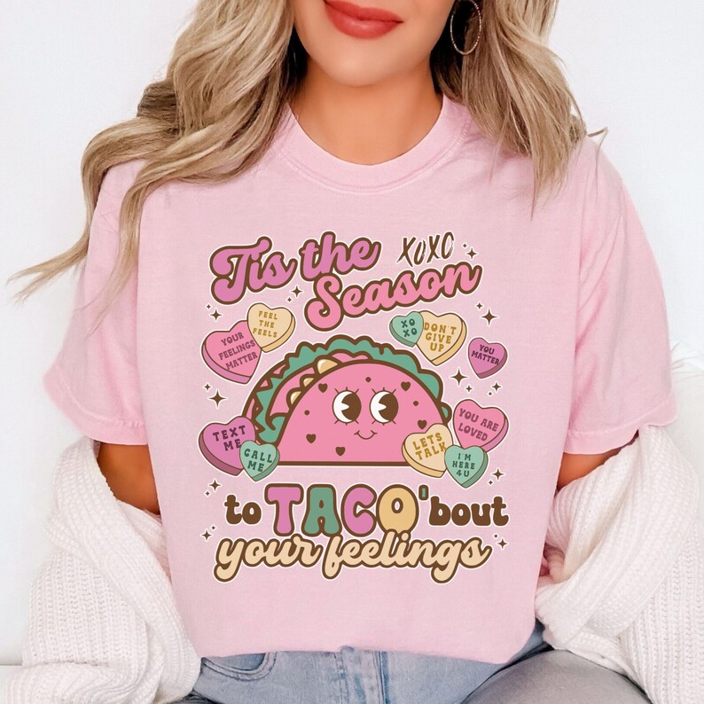 May include: Pink t-shirt with a graphic of a taco with a smiling face. The taco is surrounded by candy hearts with messages like "Feel the Feels", "You Matter", and "Let's Talk". The text on the shirt reads "Tis the Season to Taco'bout your feelings".