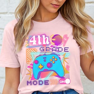 May include: A light pink t-shirt featuring a colorful graphic design. The design includes the text "4th GRADE" above a blue video game controller. Below the controller, the text "MODE ON" is displayed.