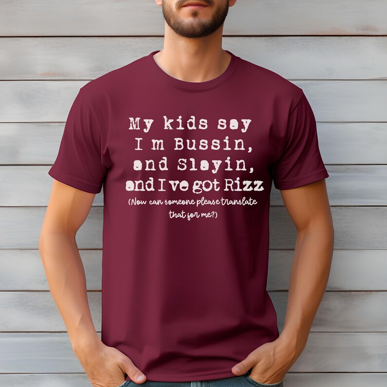 May include: A maroon t-shirt with white text. The text reads "My kids say I'm Bussin, and Slayin, and I've got Rizz (Now can someone please translate that for me?)". The shirt is a crew neck style.