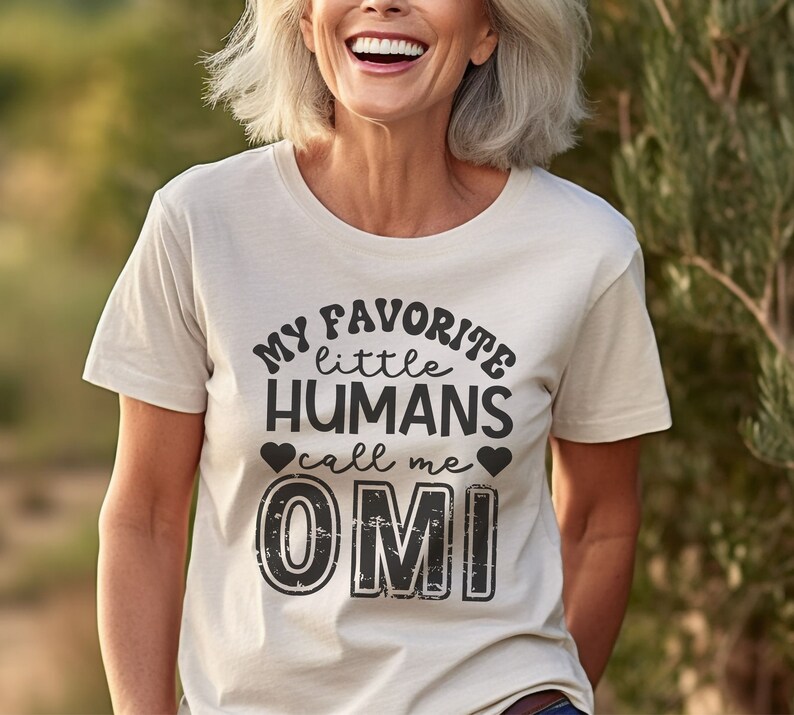 May include: A white t-shirt with black text that reads "My favorite little humans call me OMI". The text is in a distressed font and includes two small hearts.
