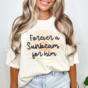 May include: A cream colored t-shirt with the text "Forever a Sunbeam for him" in black lettering. The text is surrounded by a stylized orange sun with rays.