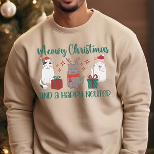 K&ouml;nnte beinhalten: Ein beiger Pullover mit einer gr&uuml;nen und wei&szlig;en Grafik von drei Katzen, die Weihnachtsm&uuml;tzen tragen. Der Text "Meowry Christmas and a Happy Neuter" ist auf dem Pullover aufgedruckt.