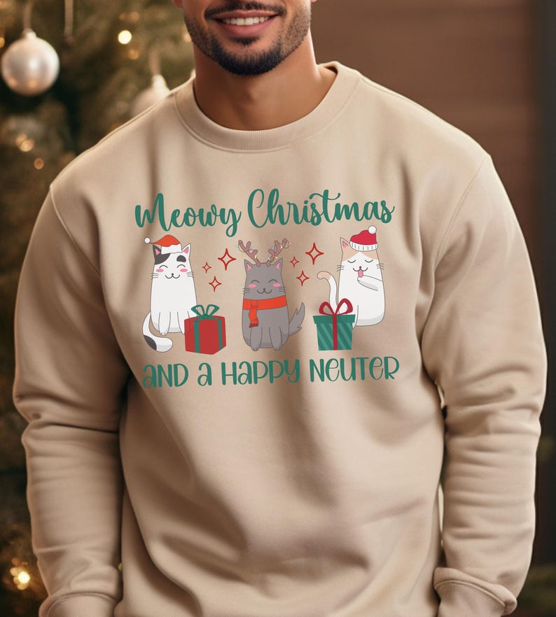 K&ouml;nnte beinhalten: Ein beiger Pullover mit einer gr&uuml;nen und wei&szlig;en Grafik von drei Katzen, die Weihnachtsm&uuml;tzen tragen. Der Text "Meowry Christmas and a Happy Neuter" ist auf dem Pullover aufgedruckt.