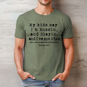 May include: Olive green t-shirt with the text "My kids say I'm Bussin, and Slayin, and I've got Rizz (Now can someone please translate that for me?)" printed in black. The shirt is short-sleeved.