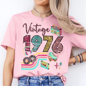 May include: Pink t-shirt with a retro design featuring the word "Vintage" above the number "1976". The design includes a boombox, roller skate, cassette tape, and records. The shirt has a crew neck.