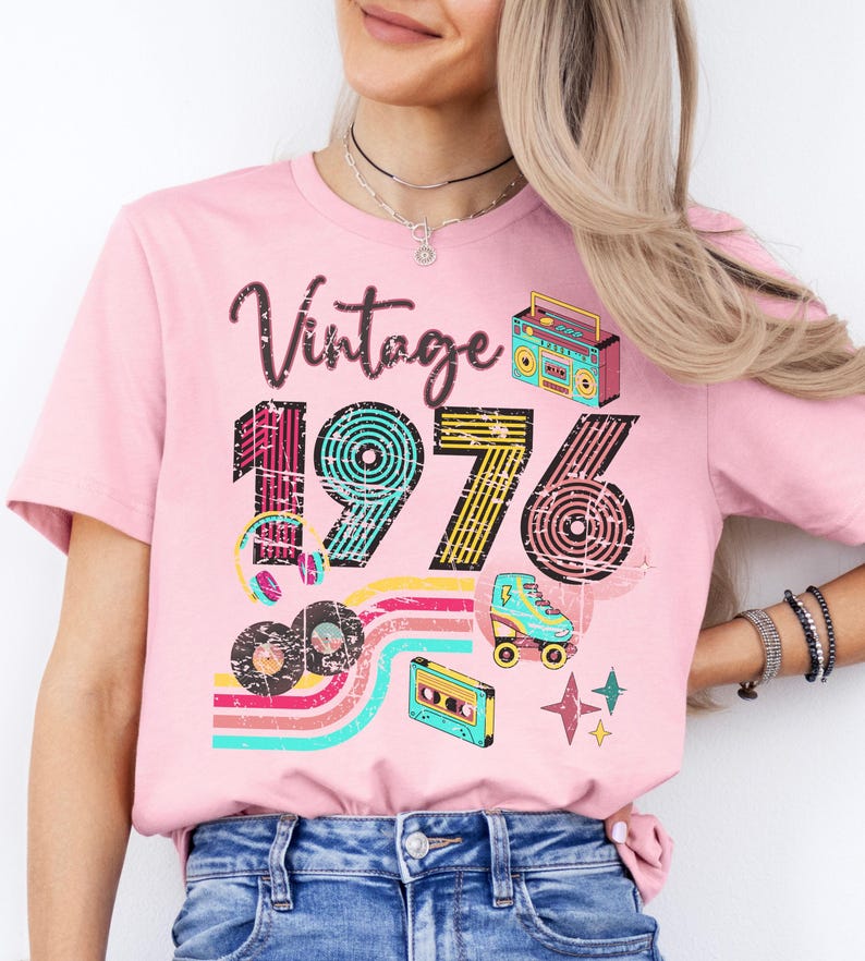 May include: Pink t-shirt with a retro design featuring the word "Vintage" above the number "1976". The design includes a boombox, roller skate, cassette tape, and records. The shirt has a crew neck.