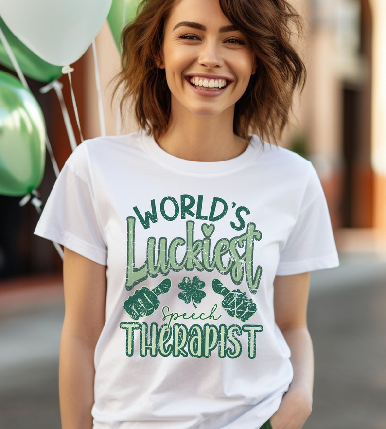 May include: White t-shirt with the text "WORLD'S Luckiest Speech Therapist" in green. The design includes a heart, a four-leaf clover, and speech bubble graphics. The shirt is worn by a person smiling, with green and white balloons in the background.
