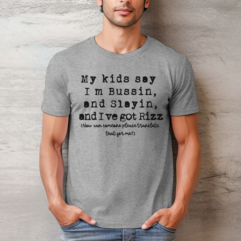 May include: A gray t-shirt with black text that reads "My kids say I'm Bussin, and Slayin, and I've got Rizz (Now can someone please translate that for me?)". The shirt is short-sleeved.