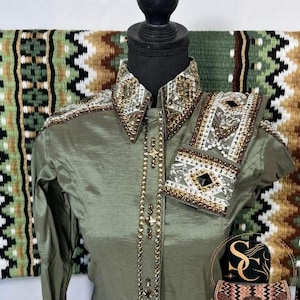 May include: An olive green show shirt with an embellished collar and cuffs, featuring gold and silver detailing. A long beaded necklace with cross pendants hangs down the front. The shirt is displayed against a green, brown, and white patterned textile.