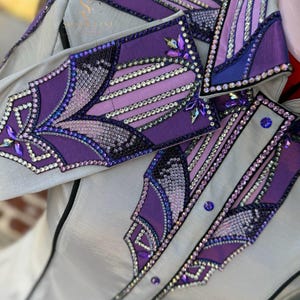 Può includere: Una camicia grigio chiaro con intricati abbellimenti viola e cristallo. La camicia presenta un colletto e polsini dettagliati con motivi geometrici, cristalli scintillanti e un bordo nero. Il design incorpora varie tonalità di viola e cristalli iridescenti.