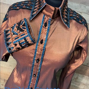 Può includere: Una camicia a maniche lunghe color rame con chiusura a bottoni, colletto a punta e intricati decori di cristalli e borchie geometriche blu, nere e color rame su colletto, spalle, polsini e sull'abbottonatura frontale.