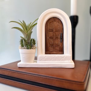 May include: A white arched structure featuring a dark brown wooden door with a small handle, set on a rectangular base. Next to the door is a small white pot containing a green air plant and several small green cacti with white moss.