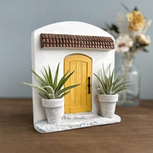 Pu&ograve; includere: Una facciata in stucco bianco in miniatura con una porta gialla, un tetto in tegole marroni e due piante in vaso. La porta ha una maniglia nera. Le piante hanno lunghe foglie verdi. Un oggetto decorativo.