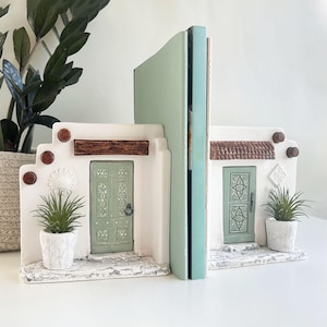 May include: White ceramic bookends shaped like miniature houses, featuring green doors, brown trim, and small potted plants. The bookends are designed to hold a book, and are placed on a white surface.