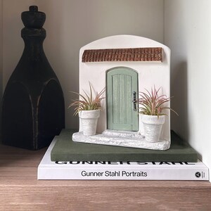Pu&ograve; includere: Una porticina decorativa con una porta verde, un telaio bianco e un tetto di tegole marroni. Due piccole piante in vaso affiancano la porta. La porta &egrave; posizionata su un blocco verde sopra un libro intitolato "Gunner Stahl Portraits". A sinistra c'&egrave; una bottiglia decorativa alta e nera.