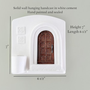 May include: White cement wall hanging with a brown wooden door, hand-painted and sealed. The piece is 7 inches high and 6.5 inches long. The text on the image says "Solid wall hanging handcast in white cement Hand painted and sealed".