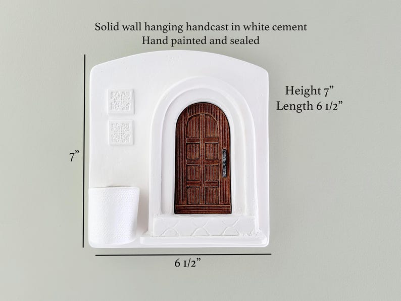 May include: White cement wall hanging with a brown wooden door, hand-painted and sealed. The piece is 7 inches high and 6.5 inches long. The text on the image says "Solid wall hanging handcast in white cement Hand painted and sealed".
