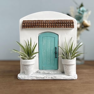 Pu&ograve; includere: Un edificio in miniatura in stucco bianco con una porta turchese, un tetto in tegole marroni e due piante in vaso. La porta ha una maniglia nera. Le piante hanno lunghe foglie verdi. &Egrave; un oggetto decorativo.
