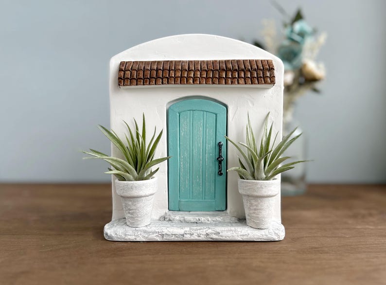 Pu&ograve; includere: Un edificio in miniatura in stucco bianco con una porta turchese, un tetto in tegole marroni e due piante in vaso. La porta ha una maniglia nera. Le piante hanno lunghe foglie verdi. &Egrave; un oggetto decorativo.