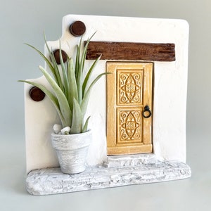 May include: A white miniature house with a carved wooden door, a brown wooden beam above the door, and a potted air plant on the side. The house is painted to look like stucco.
