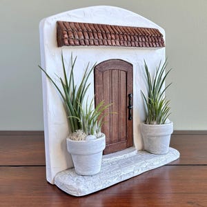 Pu&ograve; includere: Una facciata in stucco bianco in miniatura con una porta in legno marrone, un tetto in tegole di terracotta e due piante in vaso. La porta ha una maniglia nera. Le piante sono in vasi bianchi e la base &egrave; in materiale simile alla pietra.