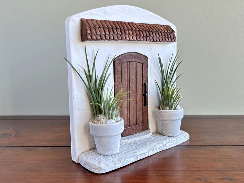 Pu&ograve; includere: Una facciata in stucco bianco in miniatura con una porta in legno marrone, un tetto in tegole di terracotta e due piante in vaso. La porta ha una maniglia nera. Le piante sono in vasi bianchi e la base &egrave; in materiale simile alla pietra.