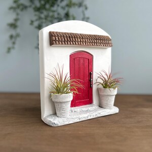 Pu&ograve; includere: Una facciata in stucco bianco in miniatura con una porta rossa, un tetto in tegole marroni e due piante in vaso. La porta ha una maniglia nera. Le piante hanno foglie verdi e rosse. La facciata &egrave; su una base bianca.
