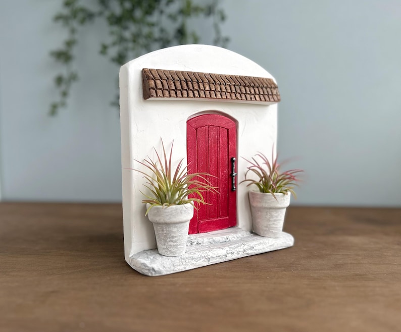 Pu&ograve; includere: Una facciata in stucco bianco in miniatura con una porta rossa, un tetto in tegole marroni e due piante in vaso. La porta ha una maniglia nera. Le piante hanno foglie verdi e rosse. La facciata &egrave; su una base bianca.